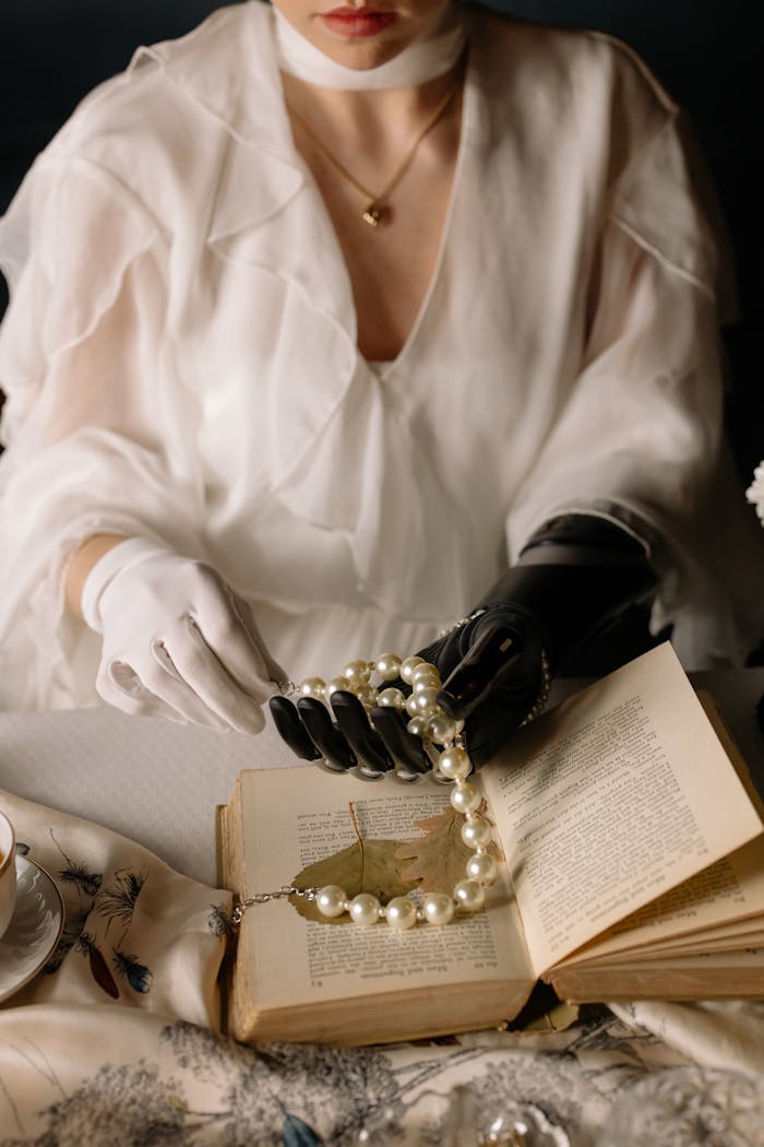 A woman with a bionic hand elegantly handles a pearl necklace over an open book.